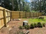 Wooden fence enclosing a grassy yard with bushes and a tree line in the background.