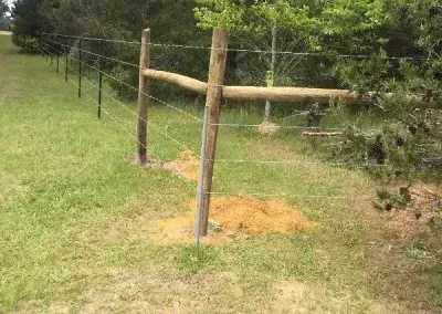A wooden fence with barbed wire in a grassy field with a tree and a dirt mound.