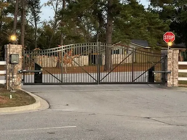 Gated entrance with brick pillars, wrought iron gate, stop sign. Light gray asphalt road, trees, and a house in the background.