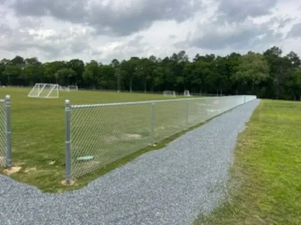 Chain-link fence and gravel path beside a soccer field with goals and trees in the background.