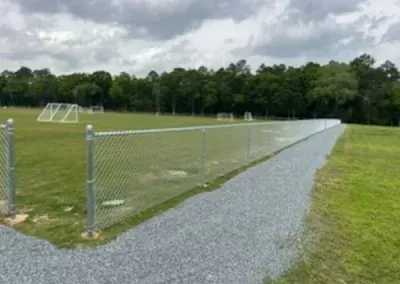 Chain-link fence and gravel path bordering a soccer field with goals and trees under cloudy sky.