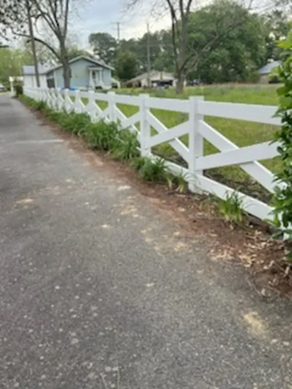 White crisscross fence along a road, with green plants in front and houses in the background.