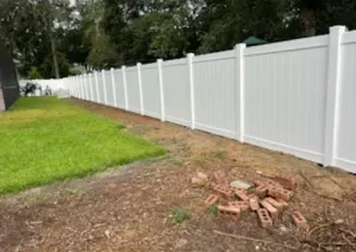 White vinyl fence bordering a grassy lawn and a dirt patch with scattered bricks.