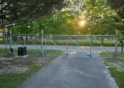 A chain-link gate blocks a paved path, with trees and a setting sun in the background.