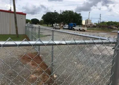 Chain link fence along a gravel road, with parked trucks and buildings in the background under a cloudy sky.
