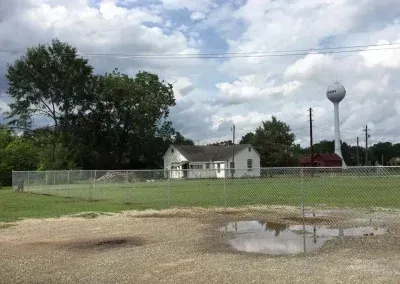 A small white building behind a chain link fence, with a water tower in the background under a cloudy sky.