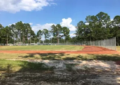 Baseball field with dirt infield, chain-link fence, trees, and blue sky with clouds.