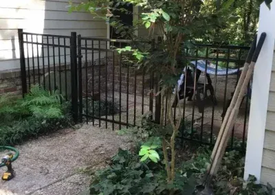 Black metal fence with gate in a yard, next to plants, gardening tools leaning against a white wall.