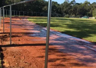 A red dirt path alongside a green grass field, with metal frames and a fence in the background.