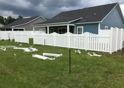 White vinyl fence under construction in a grassy yard; two houses in the background.