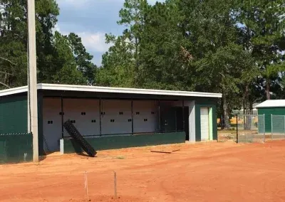 Baseball dugout with green and white trim, in a red-dirt field, with trees in the background.