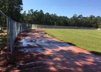 A chain-link fence bordering a wet, reddish track next to a grassy field, trees in the background.