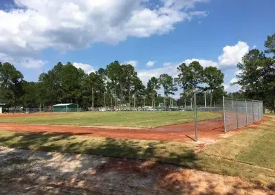 Baseball field on a sunny day. Brown dirt, green grass, chain-link fence, trees, and a blue sky with clouds.