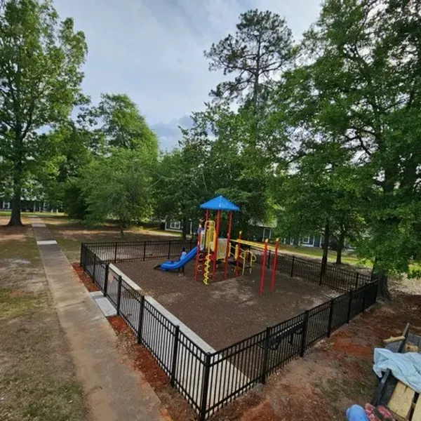 Playground enclosed by black fence, with slide, climbing bars, and blue canopy under trees.