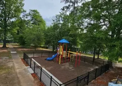 Playground enclosed by a black fence, with climbing structures, a blue slide, and surrounded by trees.