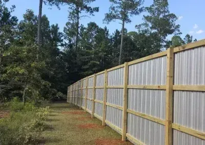 Wooden fence with metal siding curving through a wooded area, under a blue sky.