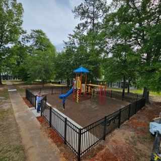 Playground enclosed by black fence, with trees in background. Person in foreground near entrance.