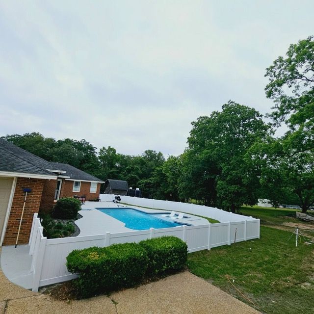 A backyard with a pool, white fence, brick house, and green trees under a cloudy sky.