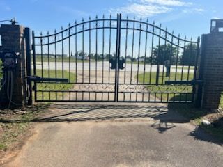 Black wrought-iron gate on a paved driveway; brick pillars on either side, sky in background.
