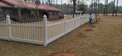 A white picket fence surrounds a brick house. A person stands next to the fence in a grassy yard.