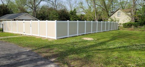 A long, tan and white vinyl fence surrounds a grassy yard, with trees and houses in the background.