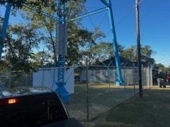 Blue amusement park ride behind a chain-link fence and a vehicle. Trees and a building are in the background.