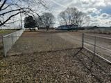 Open dirt lot with chain-link fence and gate. Bare trees and building in the background, overcast sky.
