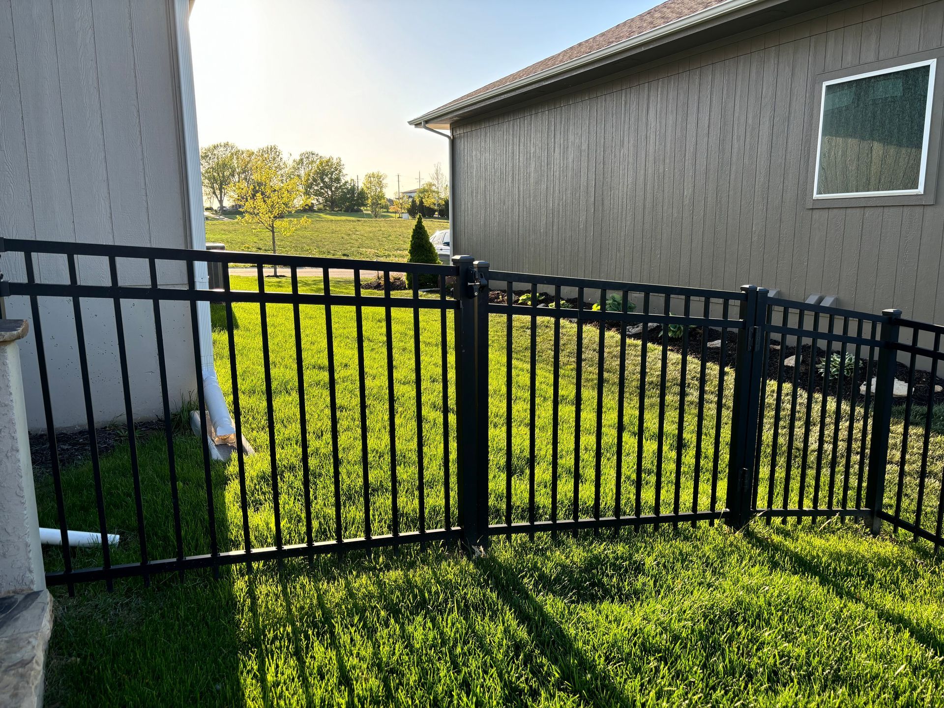 Black metal fence encloses a grassy yard between two beige houses on a sunny day.