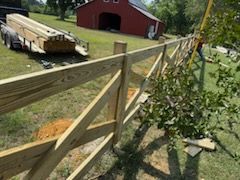 Newly constructed wooden fence with a red barn and a trailer of lumber in the background.