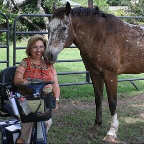 Woman in a scooter smiles next to a brown and white horse, both in a grassy field by a fence.
