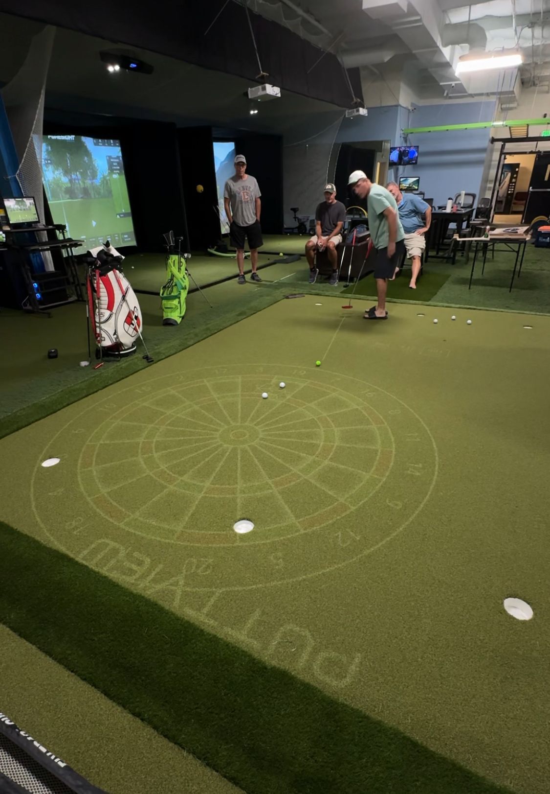 People playing indoor golf on a PuttView putting green with Foresight and Trackman Bays in background