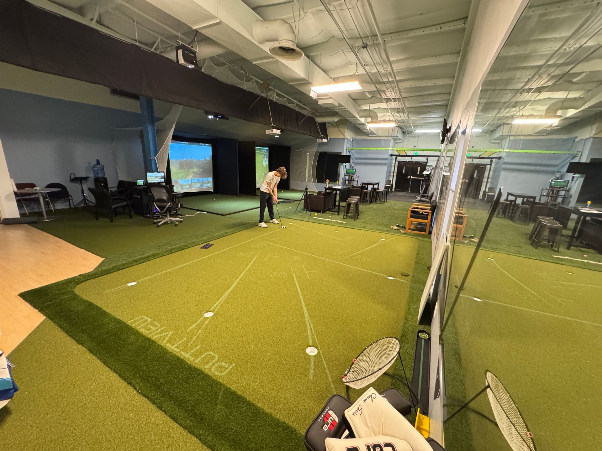 Indoor golf practice area with green turf, a golfer preparing to swing, and nets under bright ceiling lights