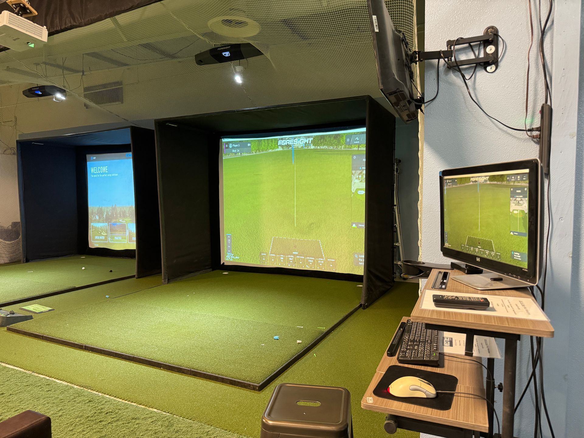 Indoor golf simulator bay with green turf, large projection screens, and a side monitor on a desk