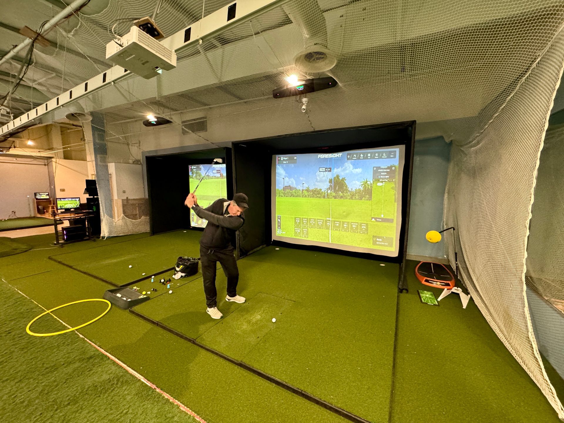 Person swinging a golf club indoors in a netted golf simulator bay with two projection screens.