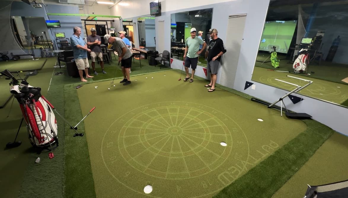 Person putting golf ball on a green indoor golf course. Others are in the background, one seated.