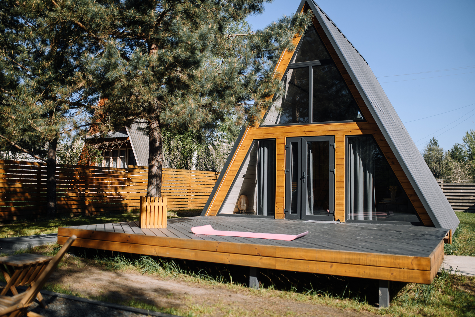 A-frame cabin with wooden deck, black roof and large windows set in a grassy area with trees.