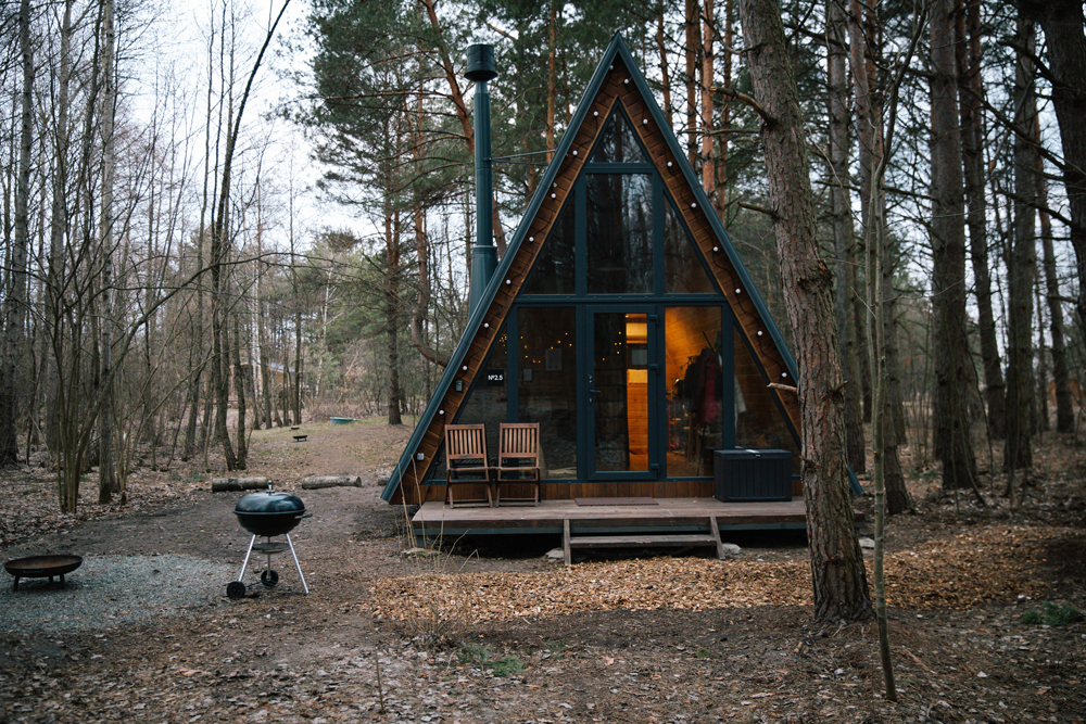 A-frame cabin nestled in a forest, with large windows, a deck, and a grill.