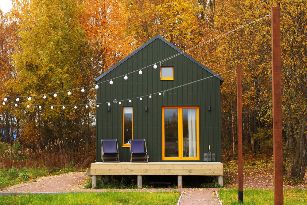 Green cabin with yellow trim and string lights on a wooden deck in front of a forest with autumn foliage.