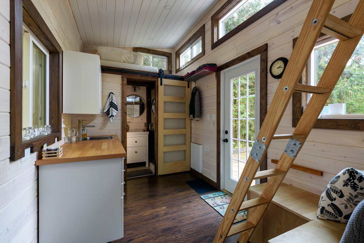 Interior of a tiny house with a kitchen, bathroom, door, and a ladder leading to a loft.