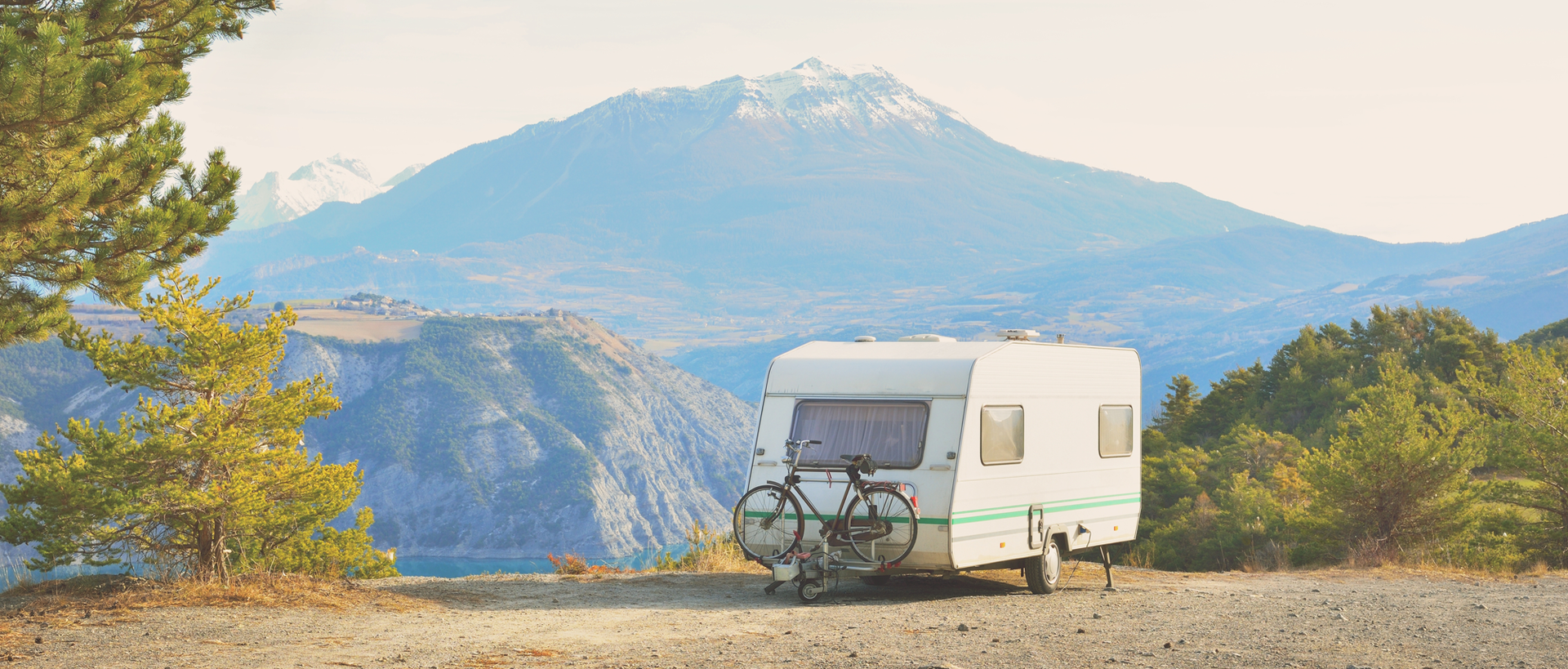 White camper trailer parked on a dirt road with mountain backdrop and bike attached.