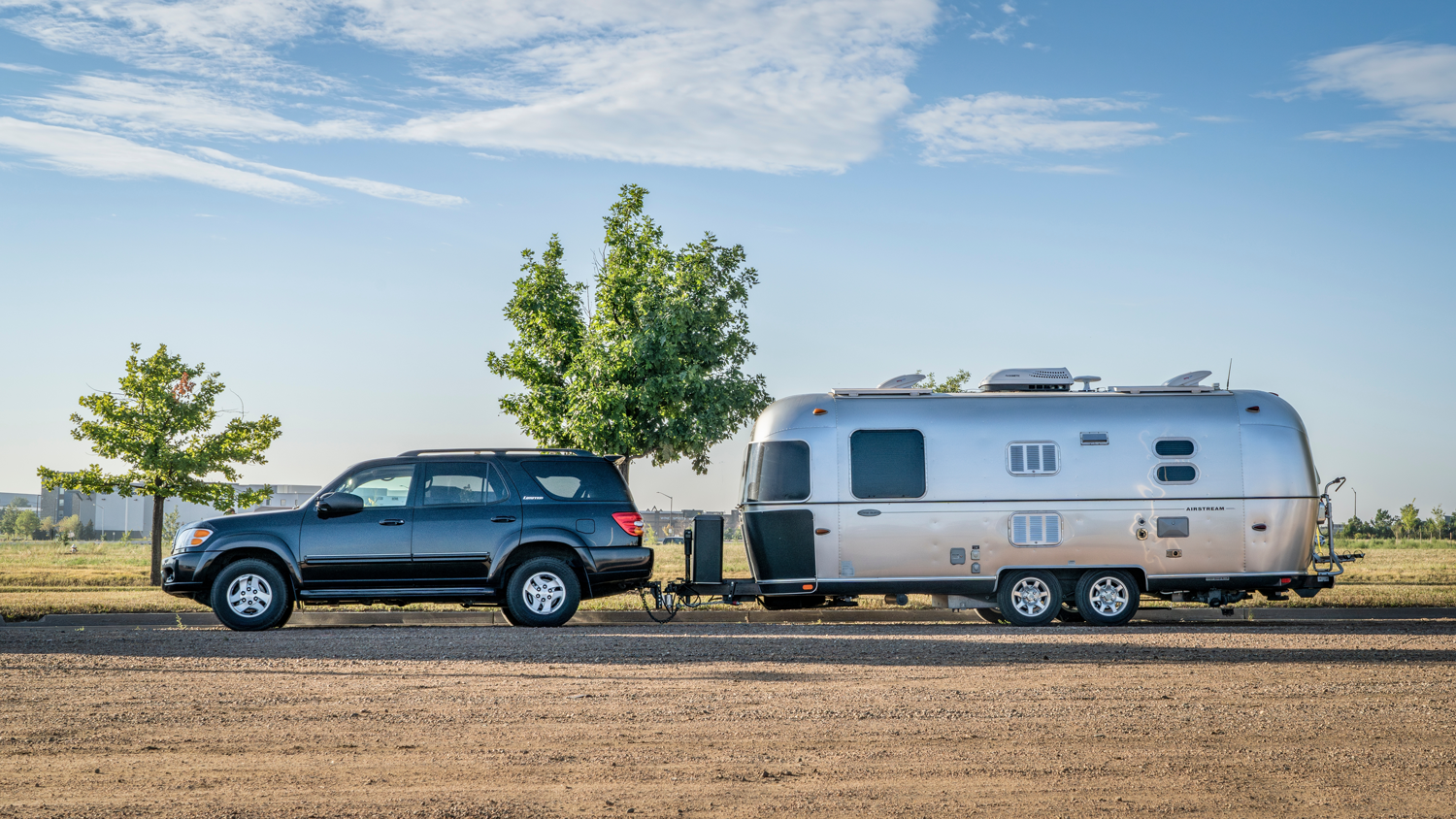 Black SUV towing a silver Airstream trailer on a dirt road, sunny day.