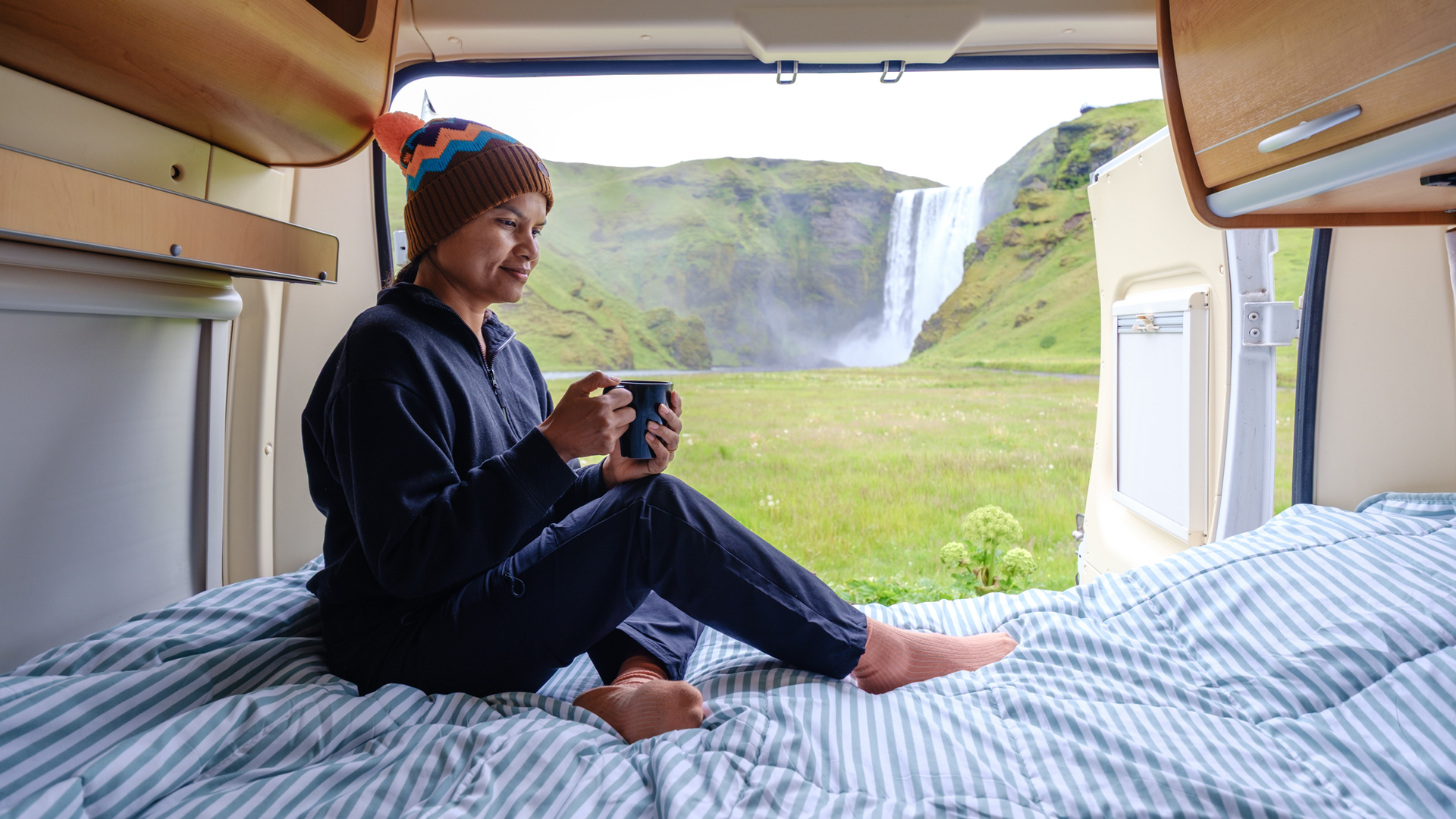 Person in pajamas with mug, sits on bed inside camper van, looking at waterfall view.