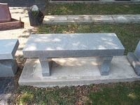 Gray stone bench on a concrete slab in a grassy area, likely a cemetery or park.