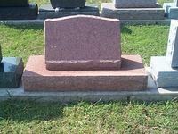 Red granite headstone on a rectangular base, set on grass.