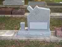 Gray tombstone with angled top and cross-like design, in a cemetery setting.