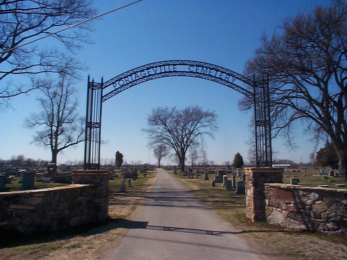 Graveled road through cemetery under a metal archway entrance. Stone pillars and bare trees. Blue sky.