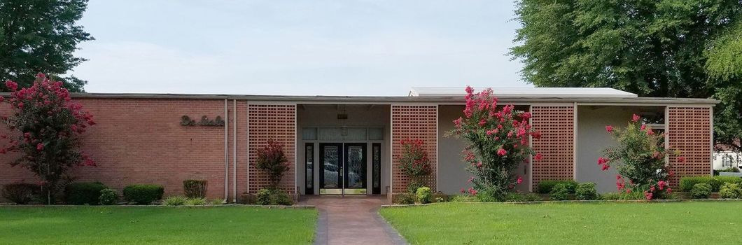 A brick building with a flat roof, surrounded by green grass and trees, and a brick pathway.