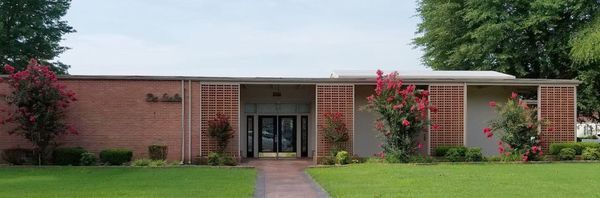Building with US flag, low-slung architecture, covered parking, and landscaping with flowers, against a sunny sky.
