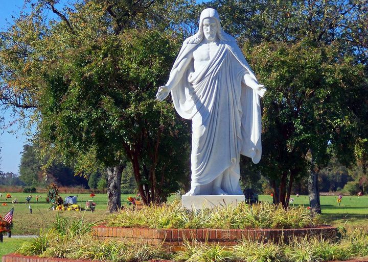 White statue of Jesus in a park, surrounded by trees and flowers under a bright sky