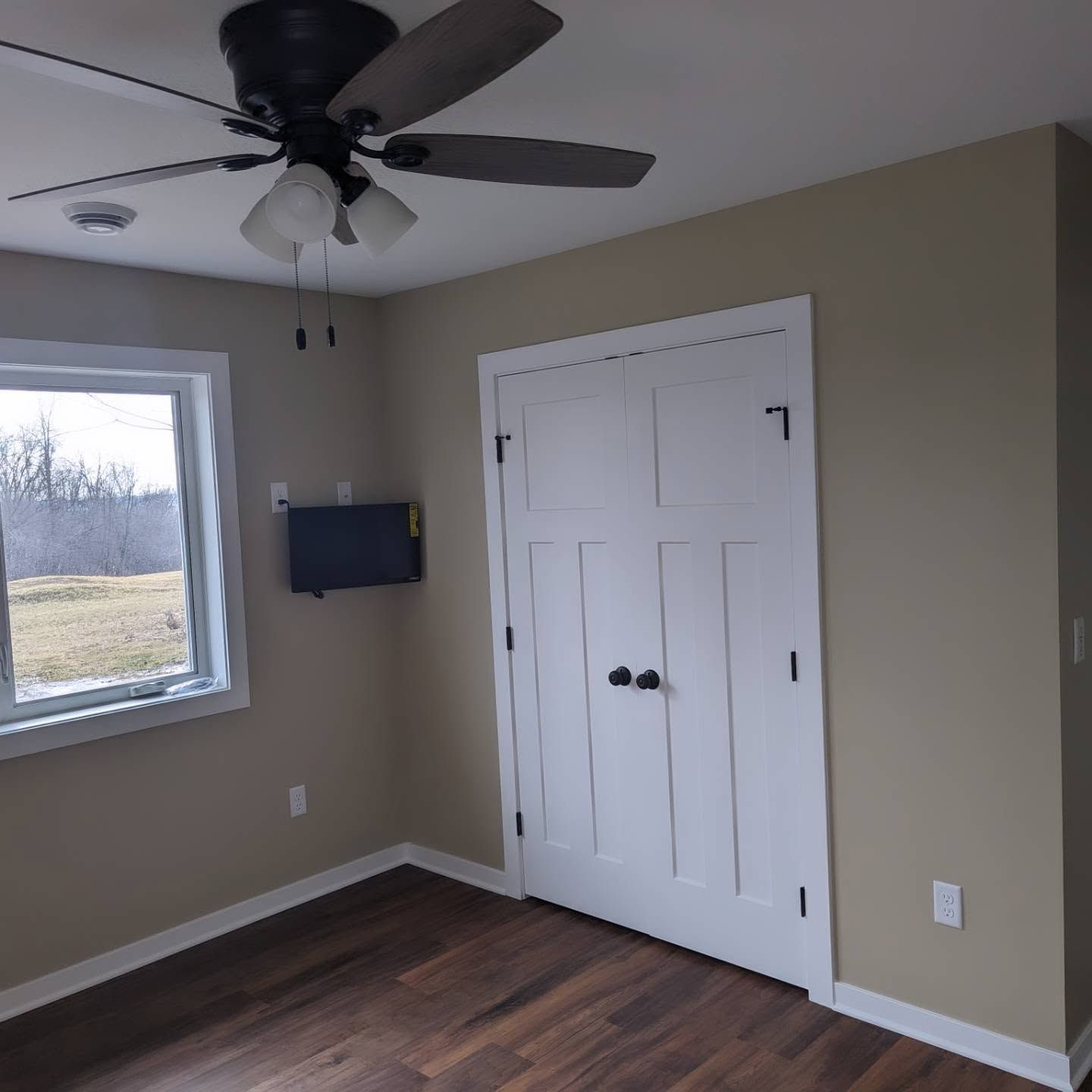 Empty bedroom with gray walls, dark wood floor, ceiling fan, window, wall TV, and white closet doors.